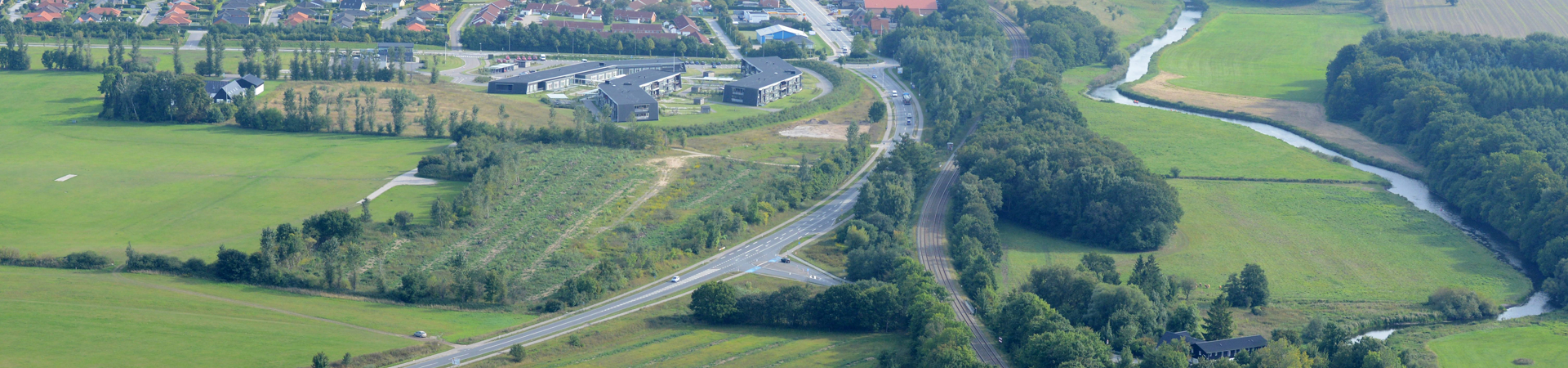 Luftfoto af et landskab med en mindre by i baggrunden og en vej og banestrækning, der løber gennem landskabet Luftfoto af et landskab med en mindre by i baggrunden og en vej og banestrækning, der løber gennem landskabet